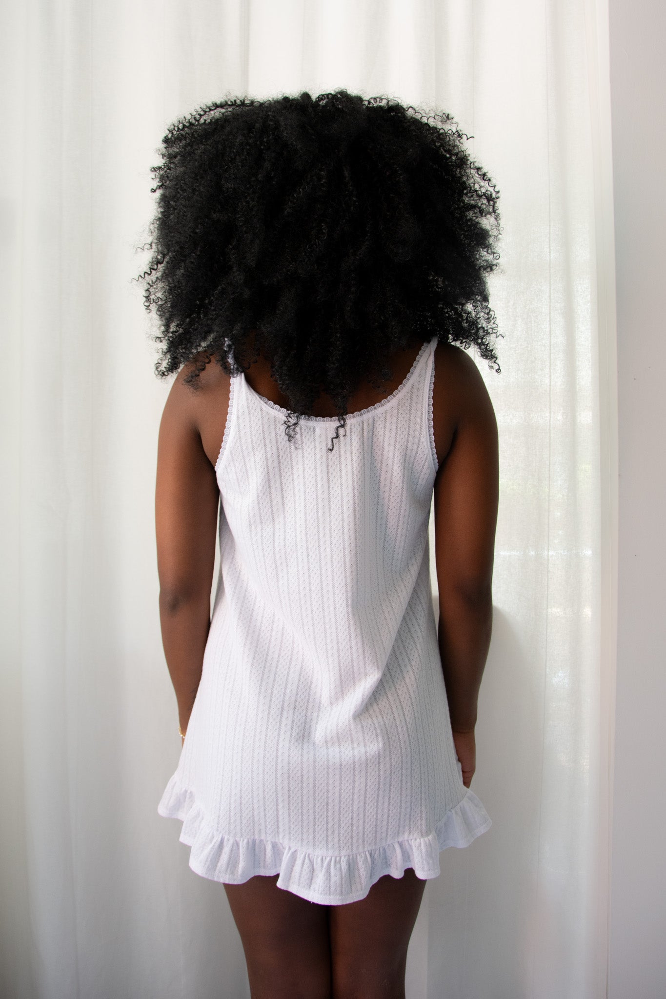 Person wearing a white sleeveless dress with ruffled hem against a white curtain background