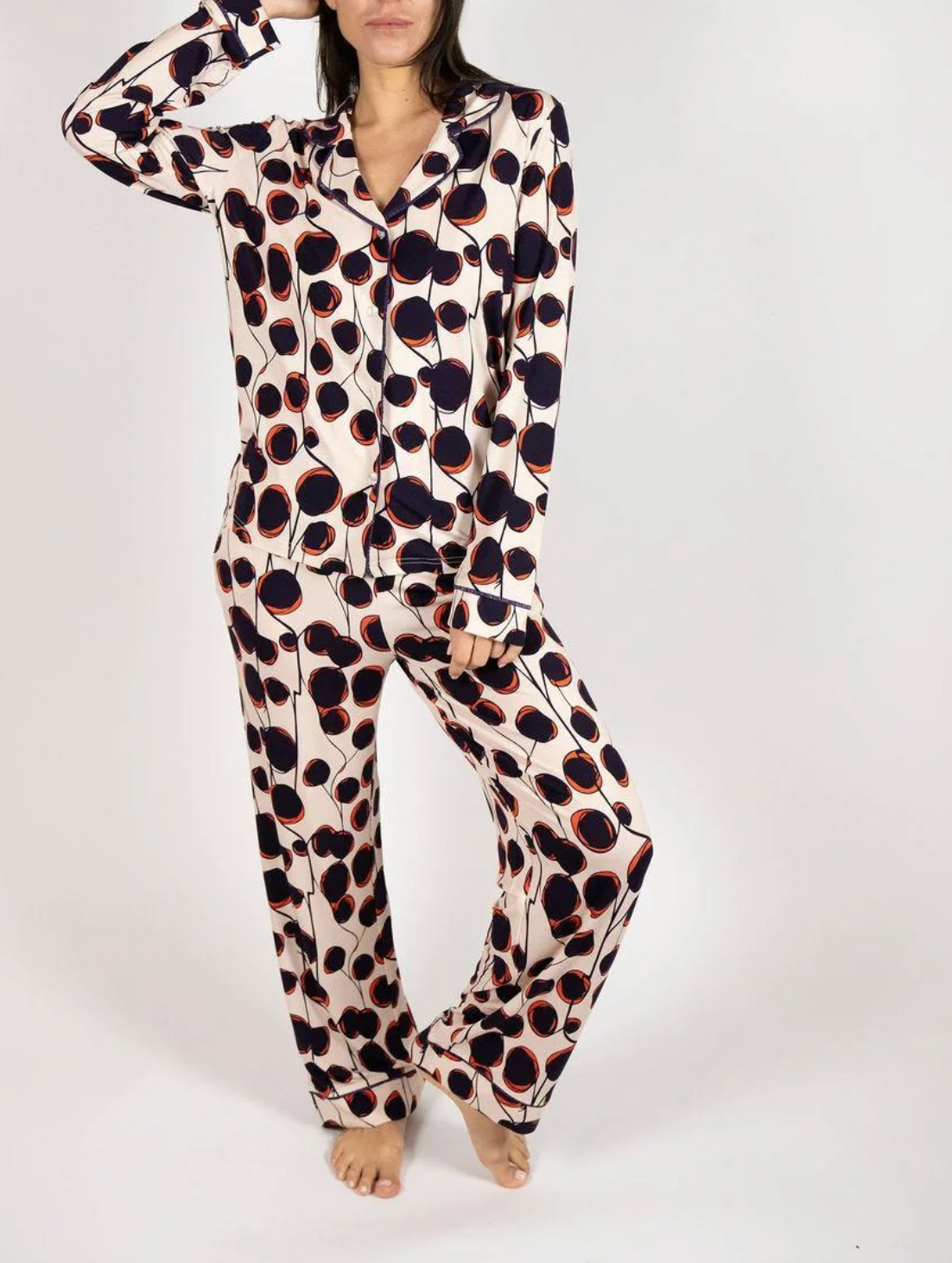 A woman modeling a matching long-sleeve pajama set with an abstract organic circle print on a cream background. The print features dark navy and black shapes outlined in orange.
