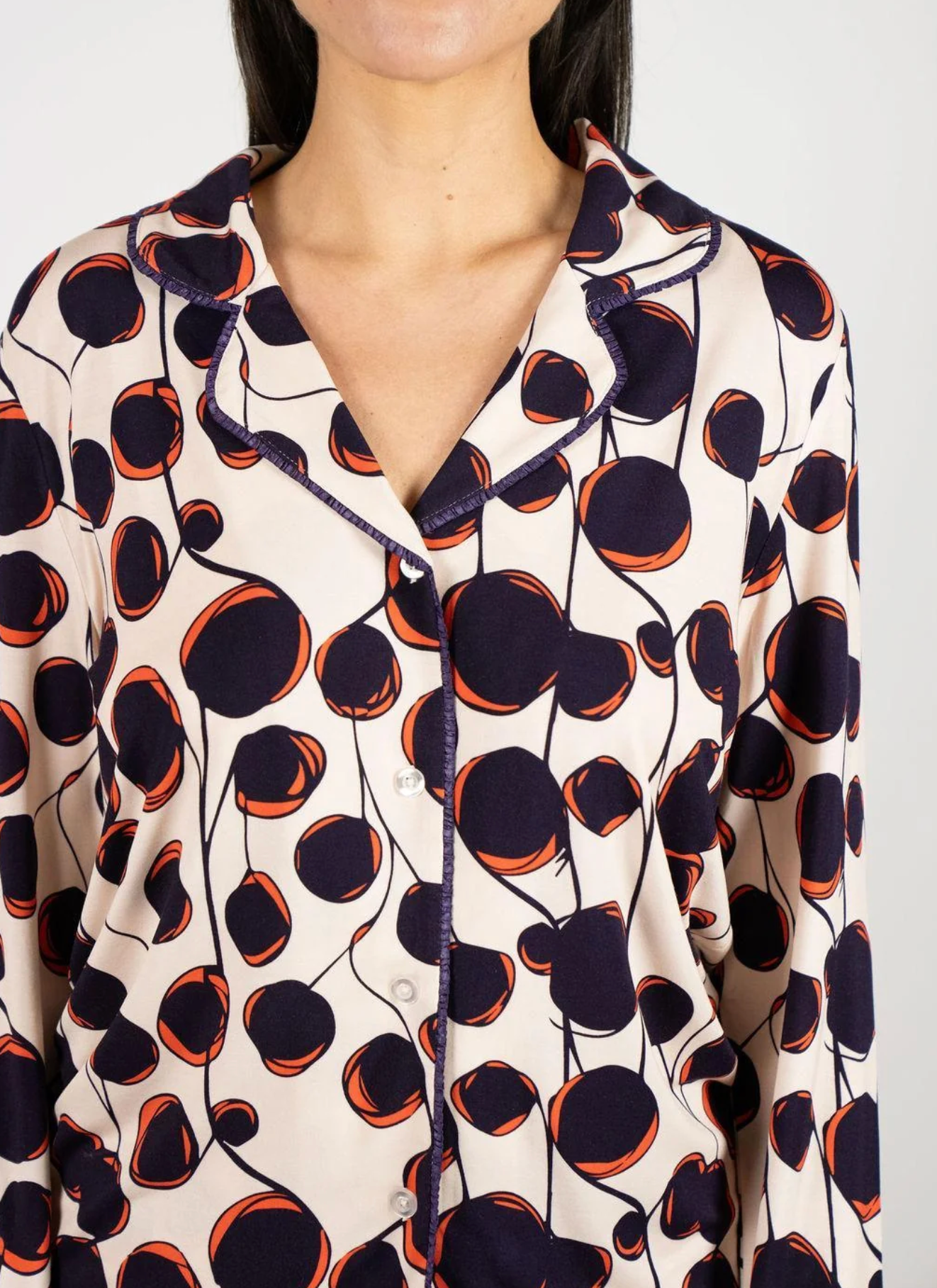 A close-up of a woman wearing a long-sleeve button-front pajama shirt with a cream base and a bold abstract print in dark navy and orange, with matching navy piping.