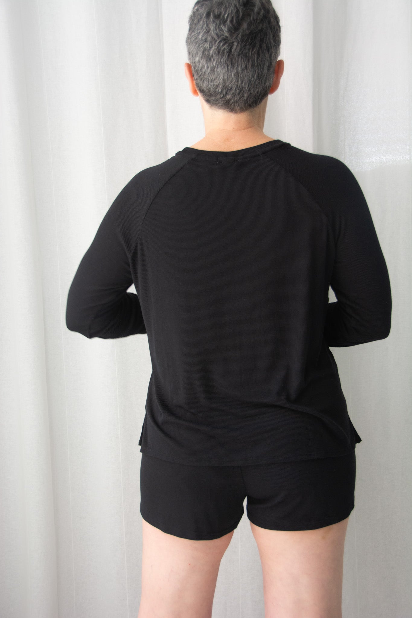 Person wearing a black long-sleeve athletic shirt and shorts against a white curtain background