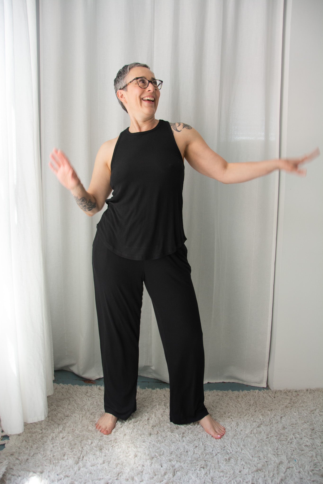 Person wearing a black outfit standing with arms outstretched in front of a white curtain.