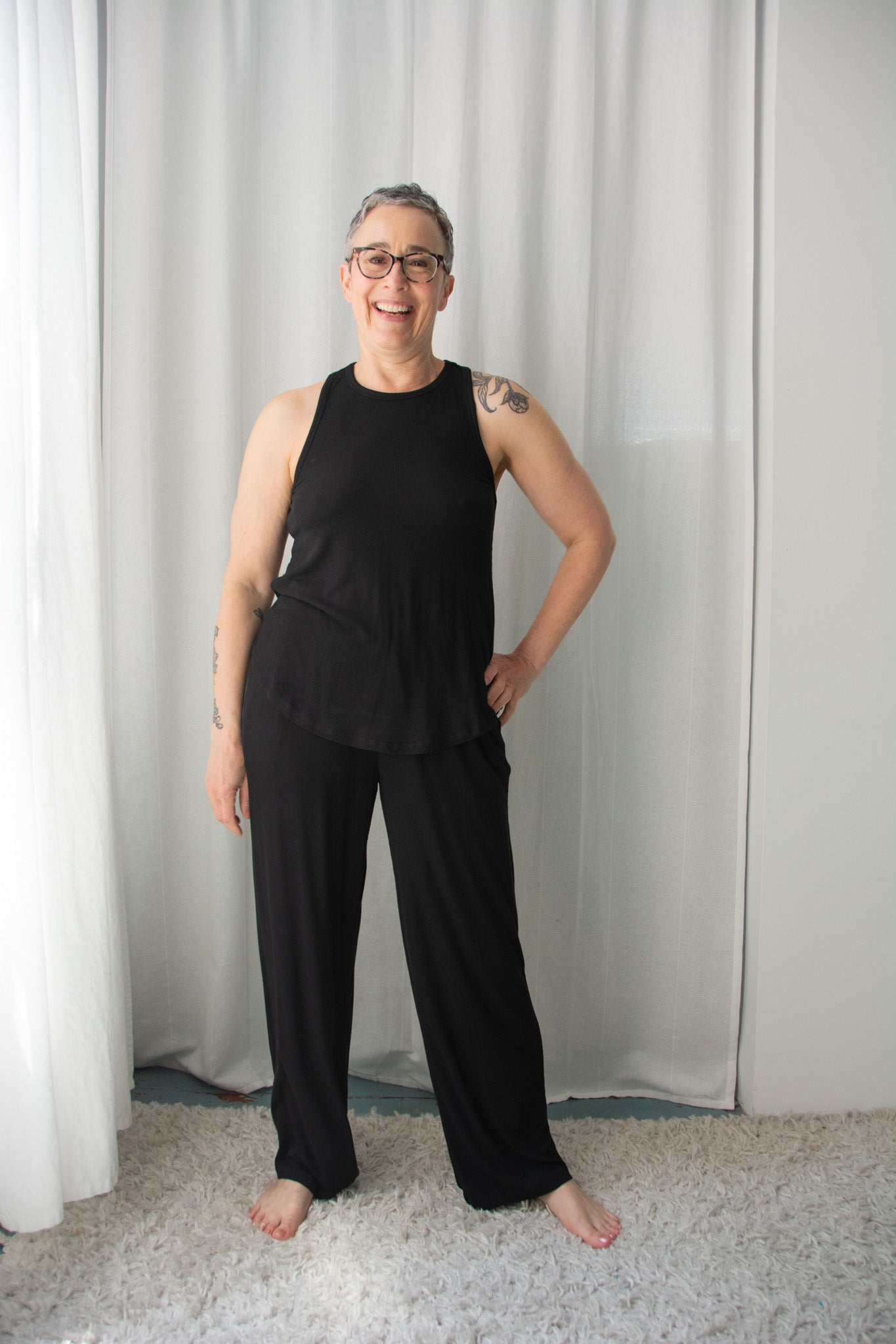 Person wearing a black sleeveless top and pants standing against a white curtain.