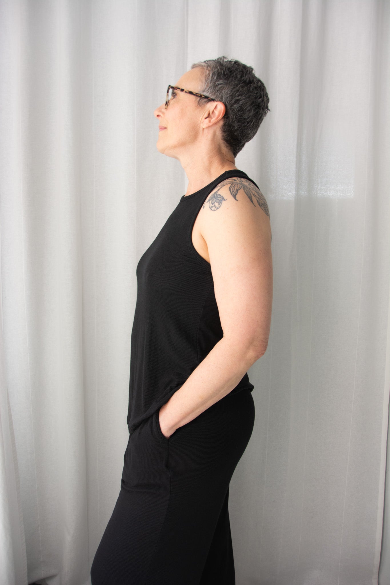 Person wearing a black sleeveless top and pants against a white curtain background