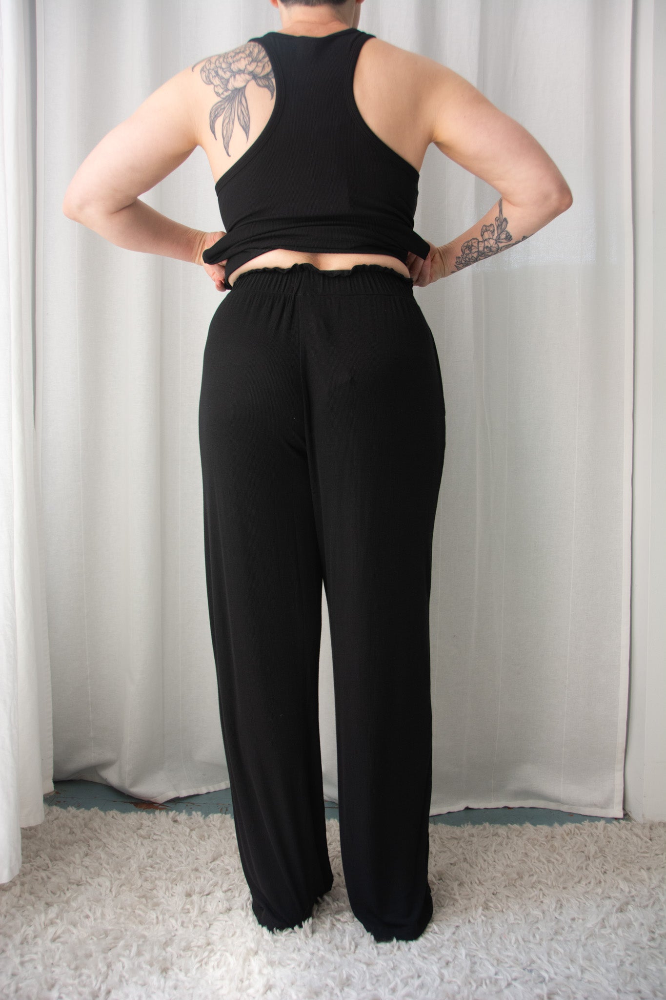 Person wearing a black tank top and wide-leg pants against a white curtain background
