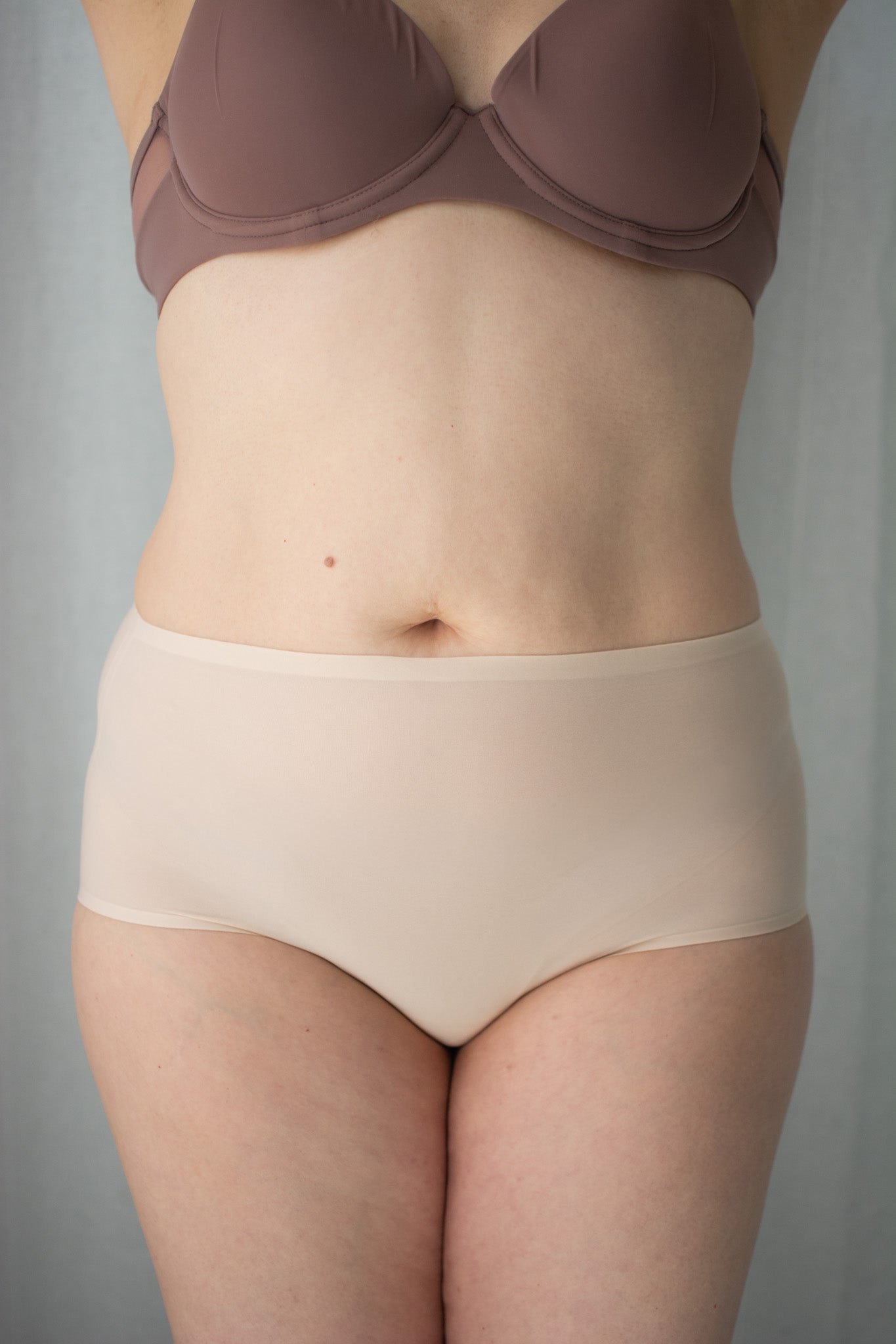 Person wearing a brown bra and beige underwear against a gray background