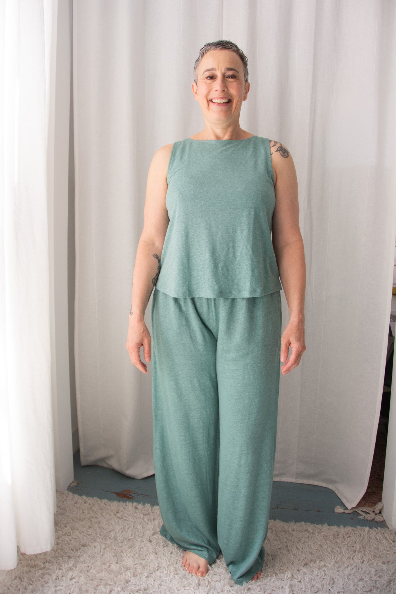 Woman wearing a green sleeveless top and pants standing in front of white curtains.