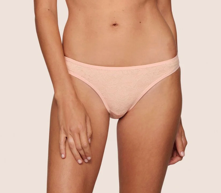 A model wearing a light peach-pink lace thong underwear against a solid light beige background.