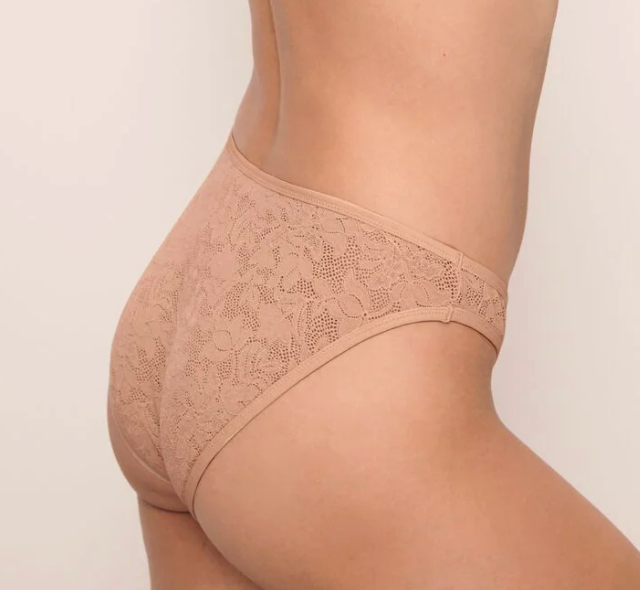 Close-up side view of a person wearing a nude lace bikini bottom against a plain light background.