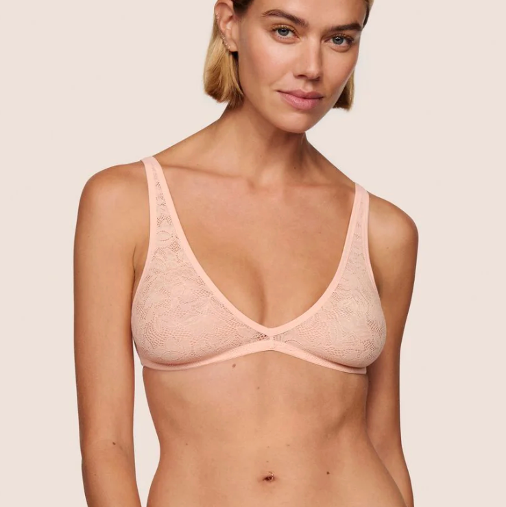 A woman modeling a light pink lace underwire bra against a solid light beige background.