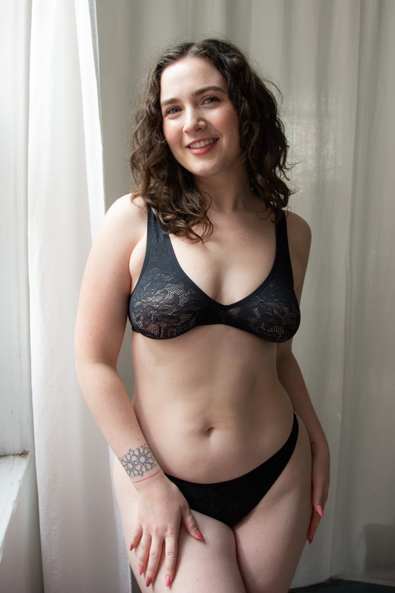 Woman wearing a black lace bra and panties set in front of white curtains.