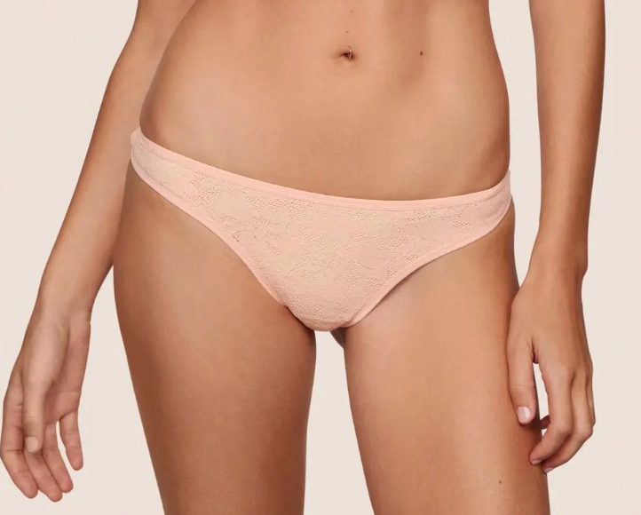 A close-up of a model wearing a light peach-pink lace thong underwear against a plain light beige background.