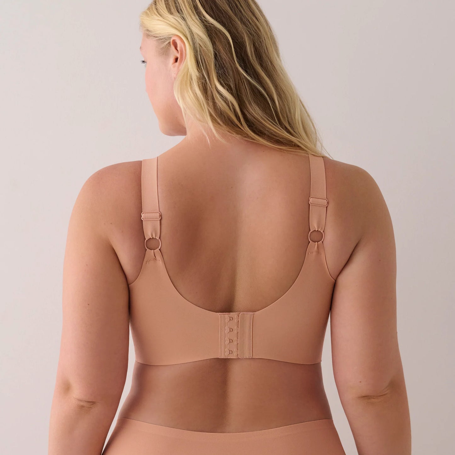 A woman shown from the back wearing a seamless, nude-beige bra with adjustable shoulder straps and a lace-up back closure.