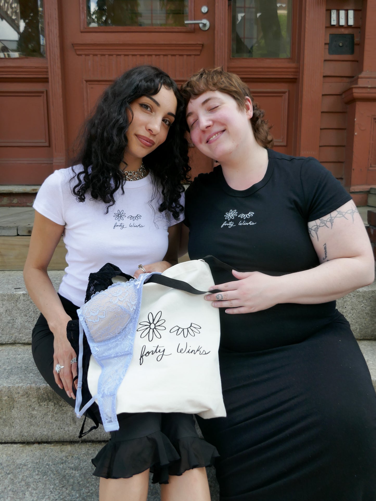 Two women holding a tote bag with a brand logo, standing on steps outside a building.
