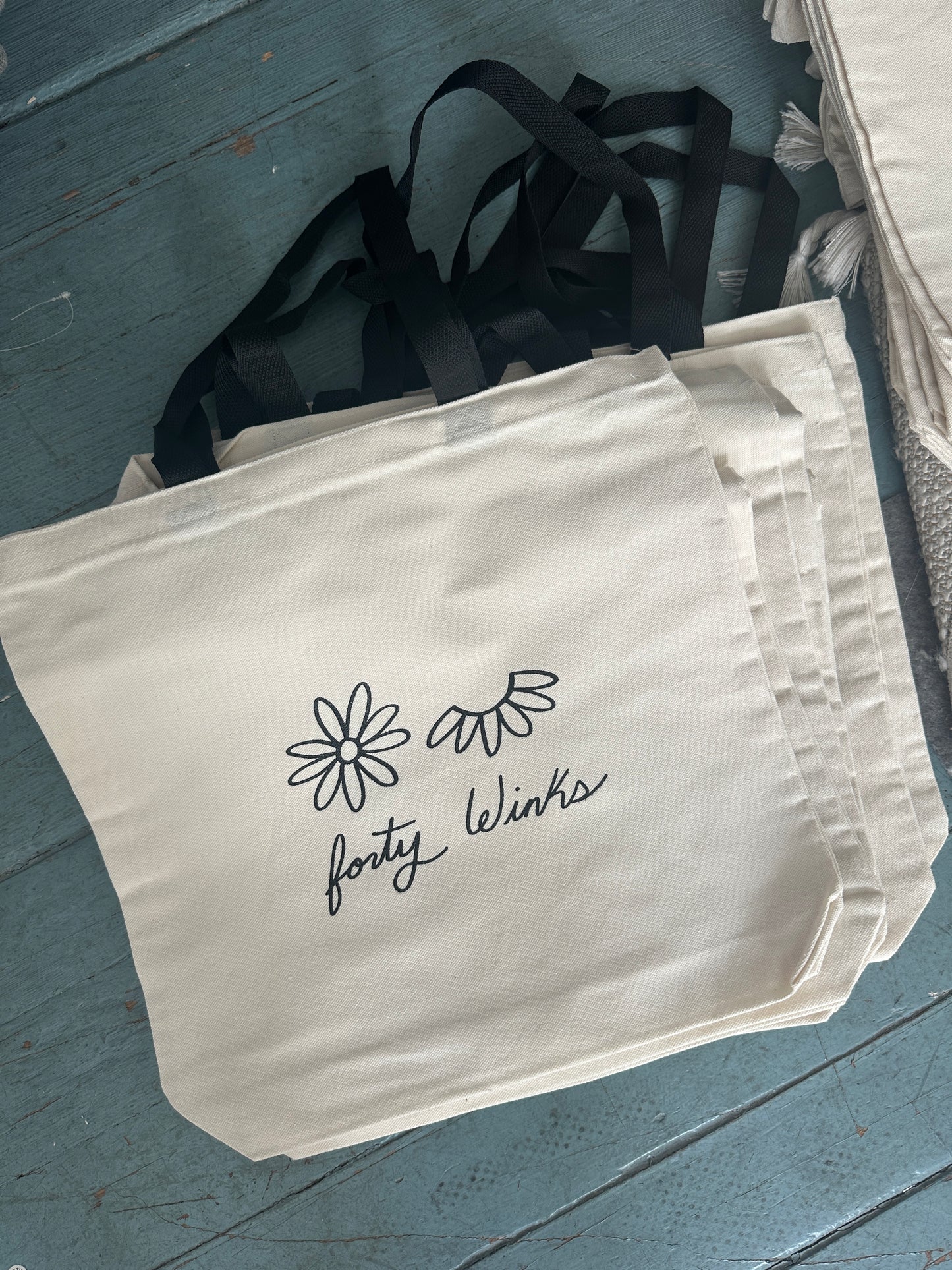 Beige tote bag with 'forty winks' text and floral designs on a blue wooden surface