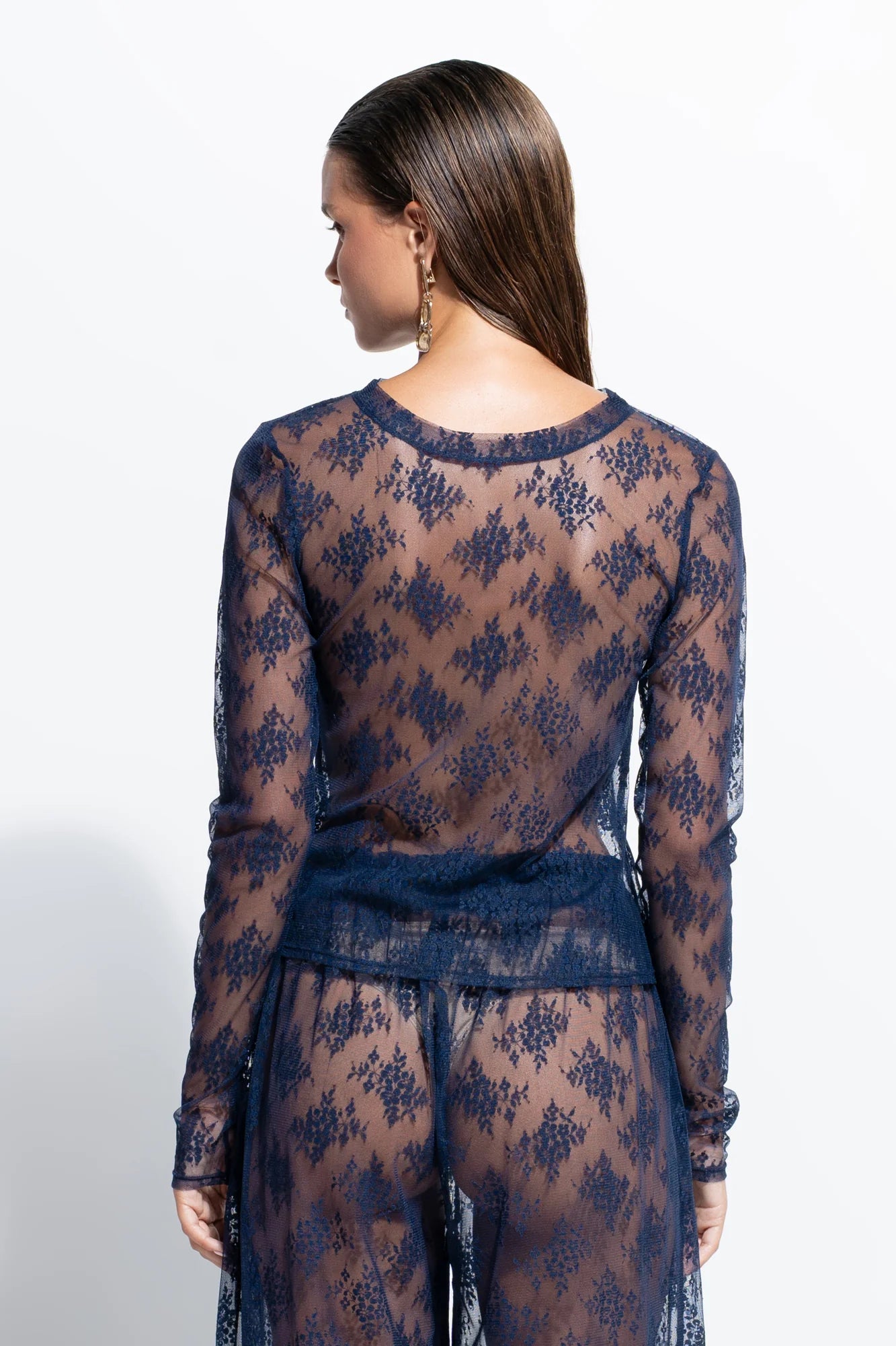 A woman shown from the back wearing a sheer navy lace long-sleeve top and matching pants against a plain white studio background.