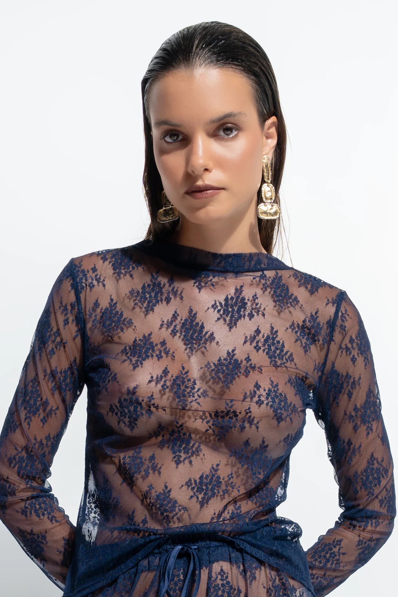 A woman modeling a sheer navy blue lace long-sleeve top with a matching waist tie, set against a plain white studio background.
