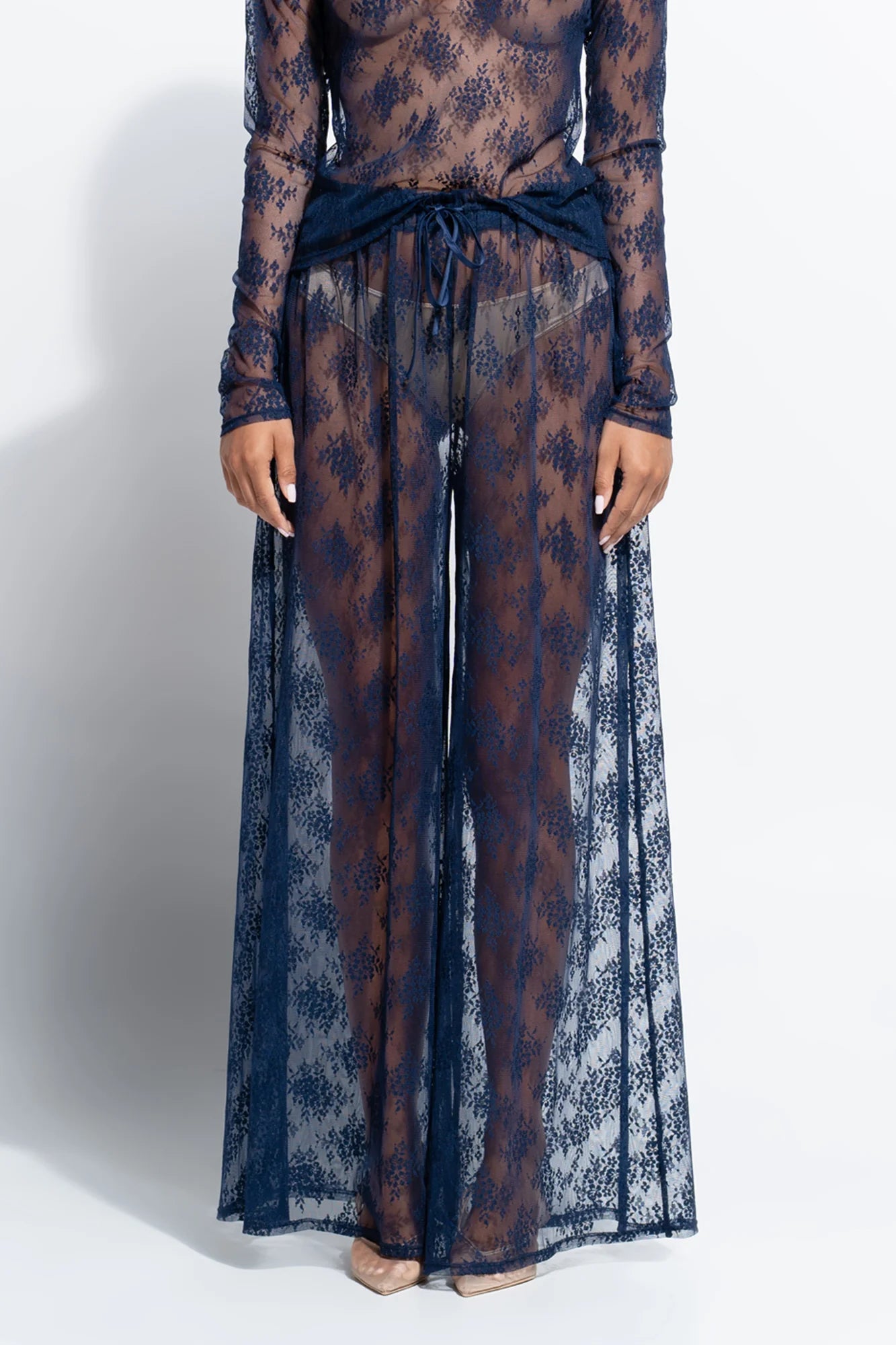 A model wearing a sheer navy blue floral lace long-sleeve top and matching wide-leg pants with a tied waist, shown against a plain white studio background.