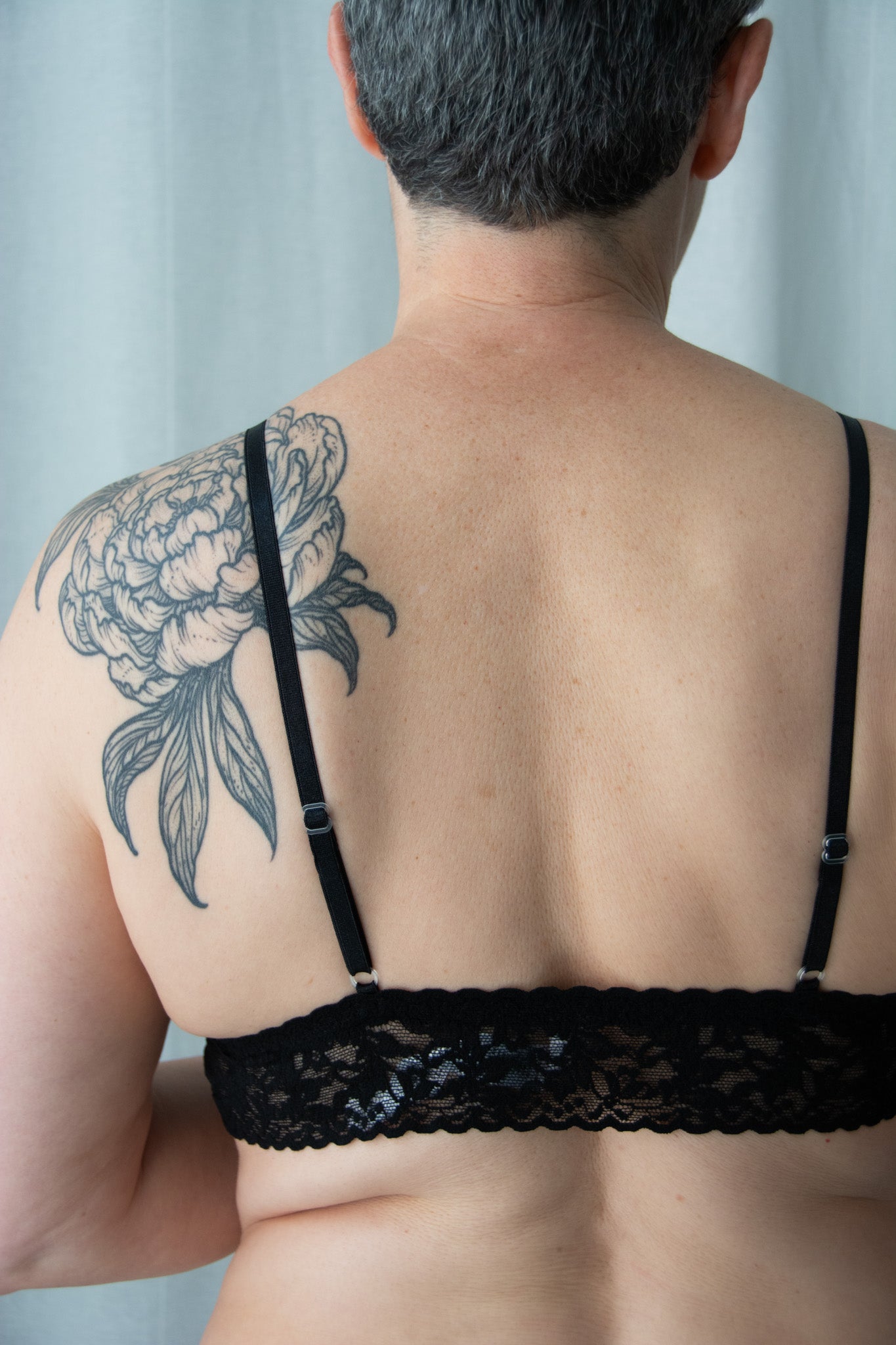 Person wearing a black lace bra with a tattoo on their shoulder against a neutral background