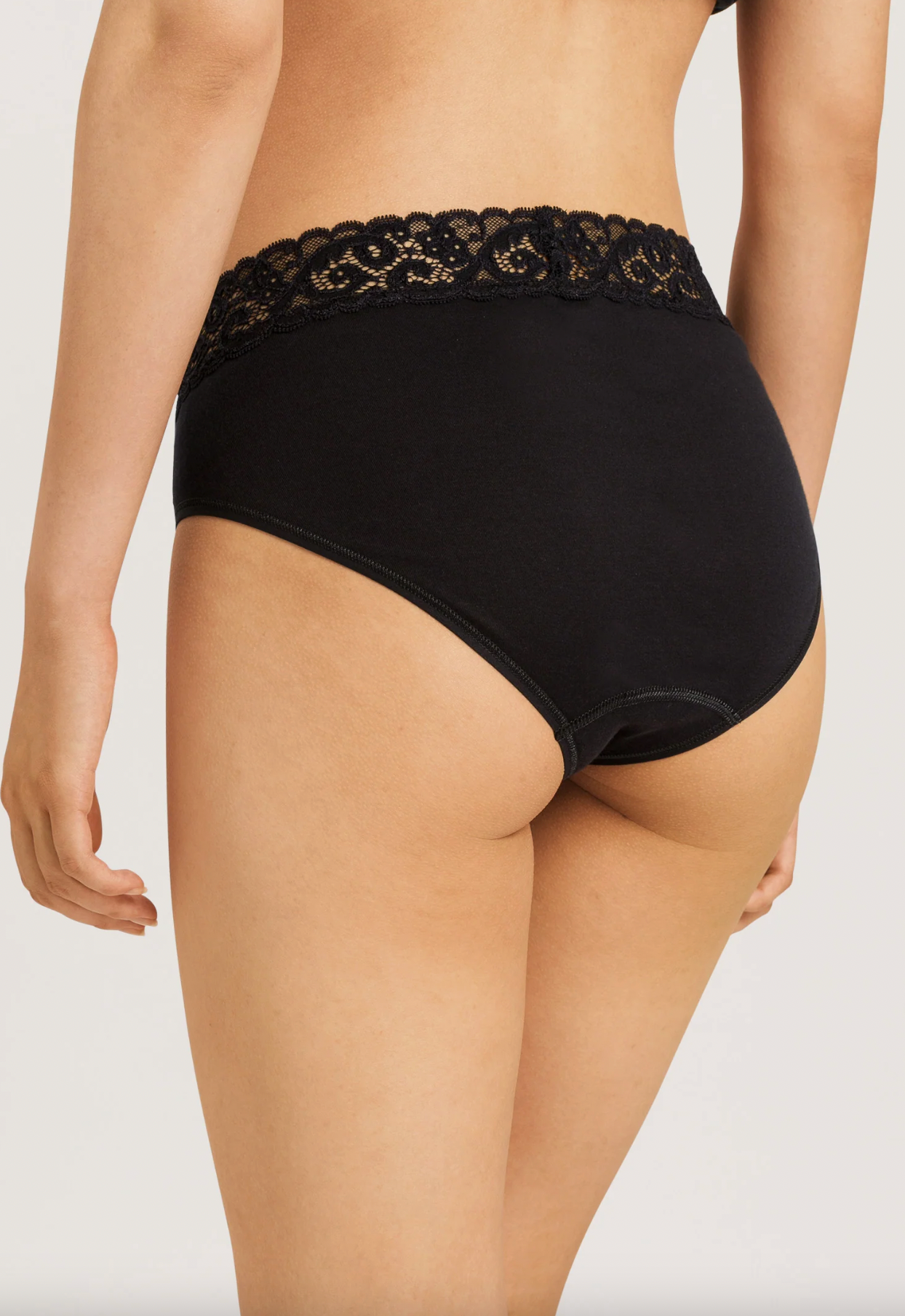 Rear view of a model wearing black high-waisted underwear with a decorative black lace waistband, set against a plain light gray studio background.