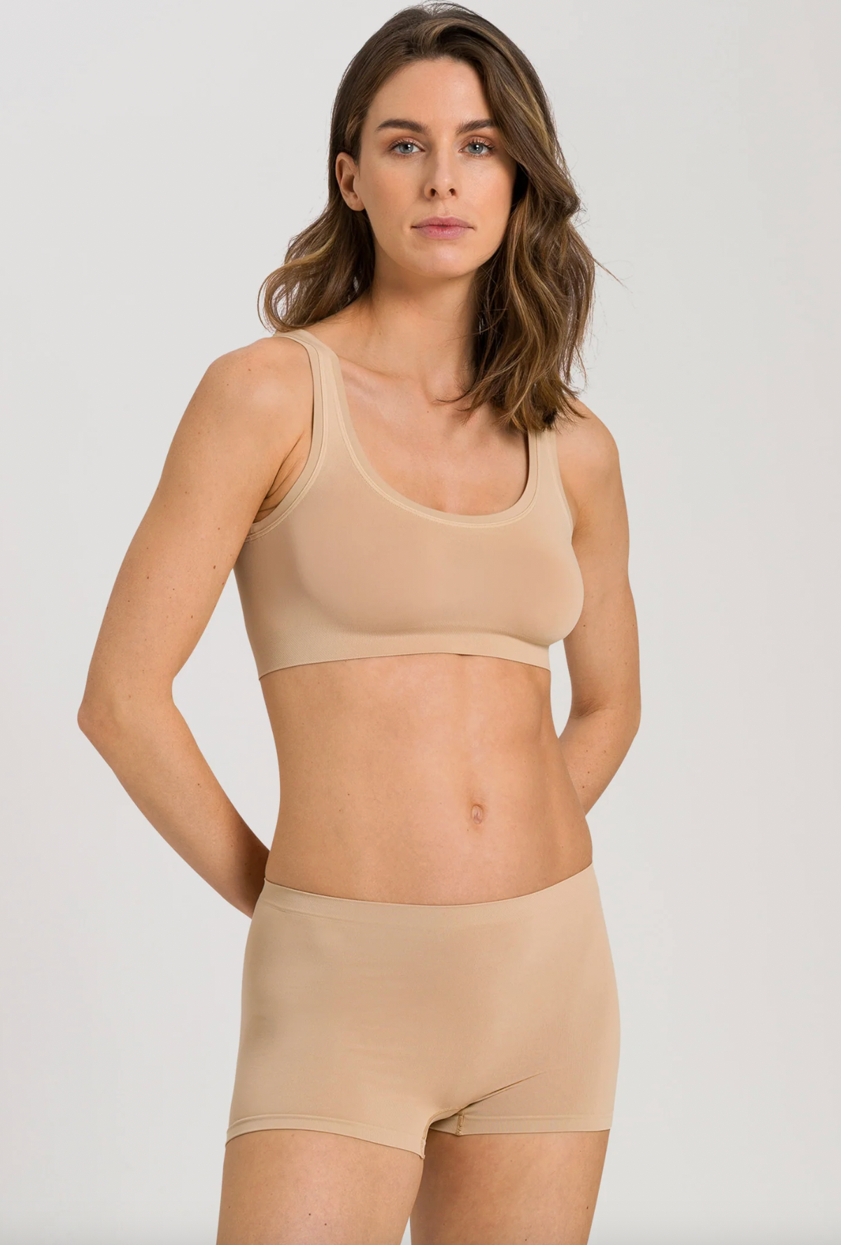 A woman modeling a matching beige two-piece lingerie set consisting of a seamless scoop-neck bralette and high-waisted briefs, shot against a light gray studio background.