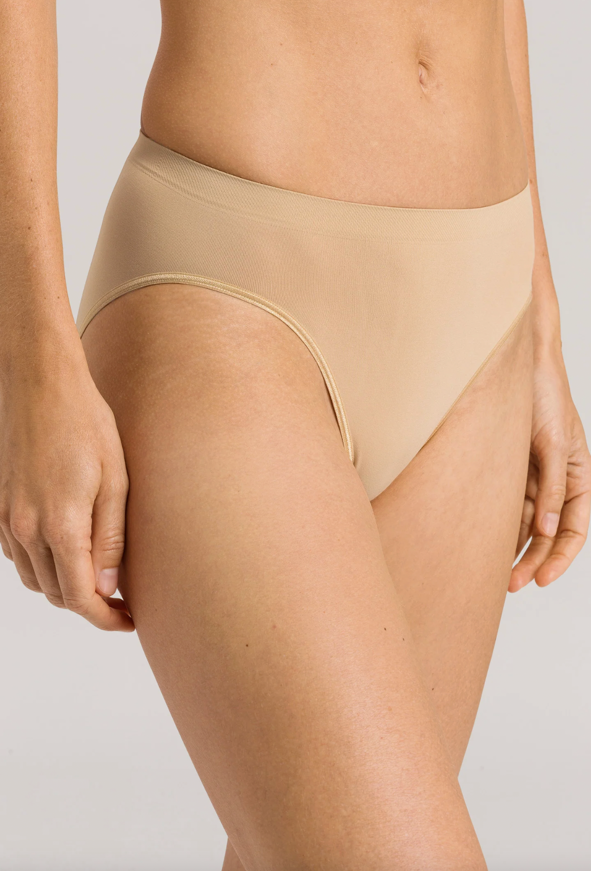 A close-up of a model wearing high-waisted, seamless beige bikini-style underwear against a light gray studio background.