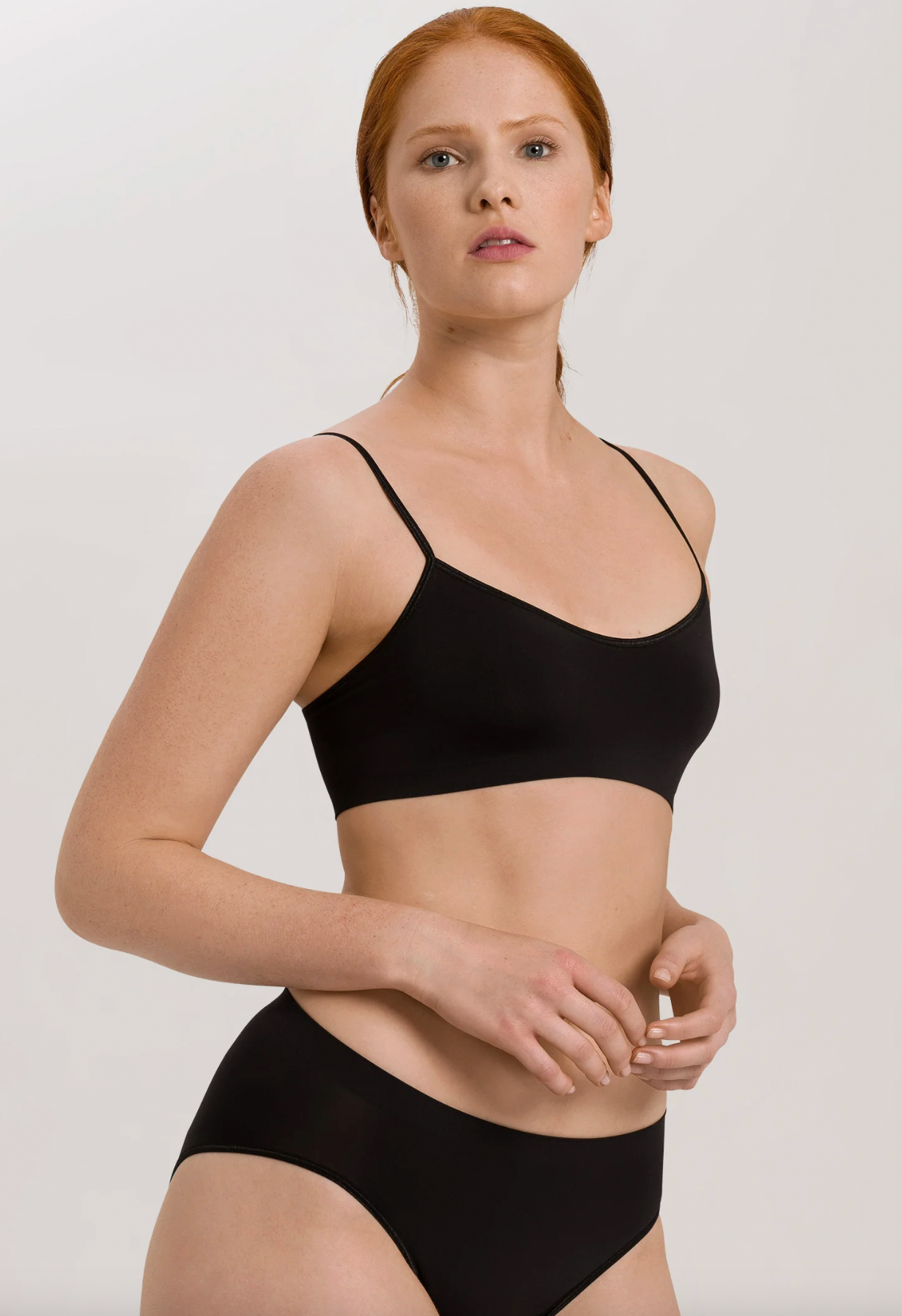 A model with red hair wearing a black bralette and matching high-waisted briefs against a light gray studio background.