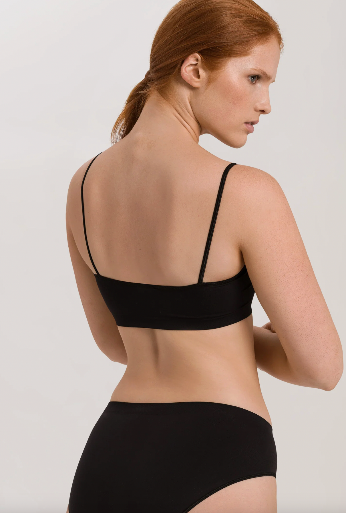 A model with red hair tied back, shown from the back wearing a black bralette and matching black underwear against a light gray studio background.