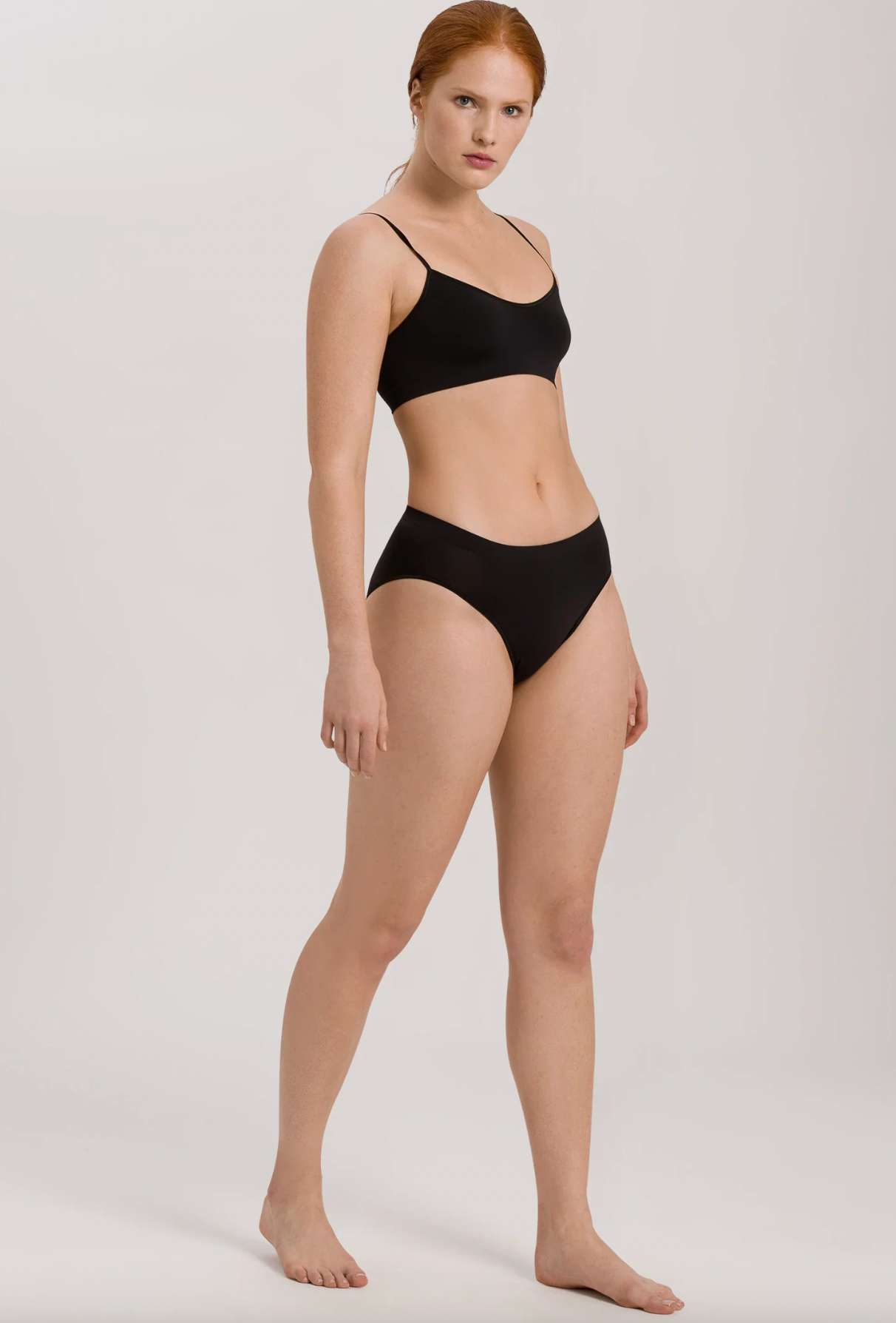 A model with red hair stands in a studio wearing a black two-piece bikini set against a light gray background.