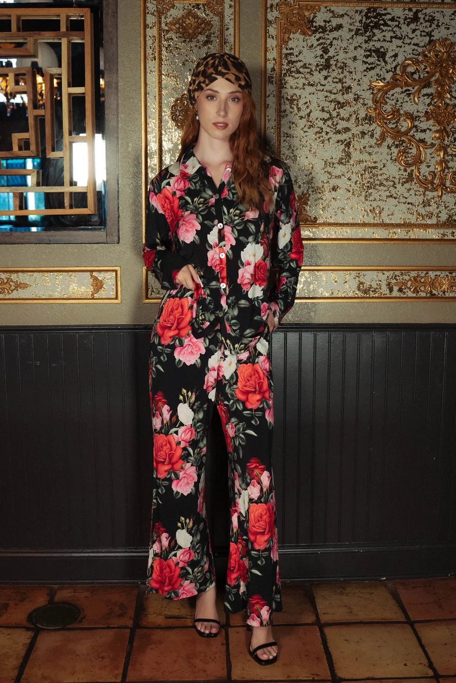 model wearing black and red floral pajamas with a button down collared top and long pants against a wall with gold details.