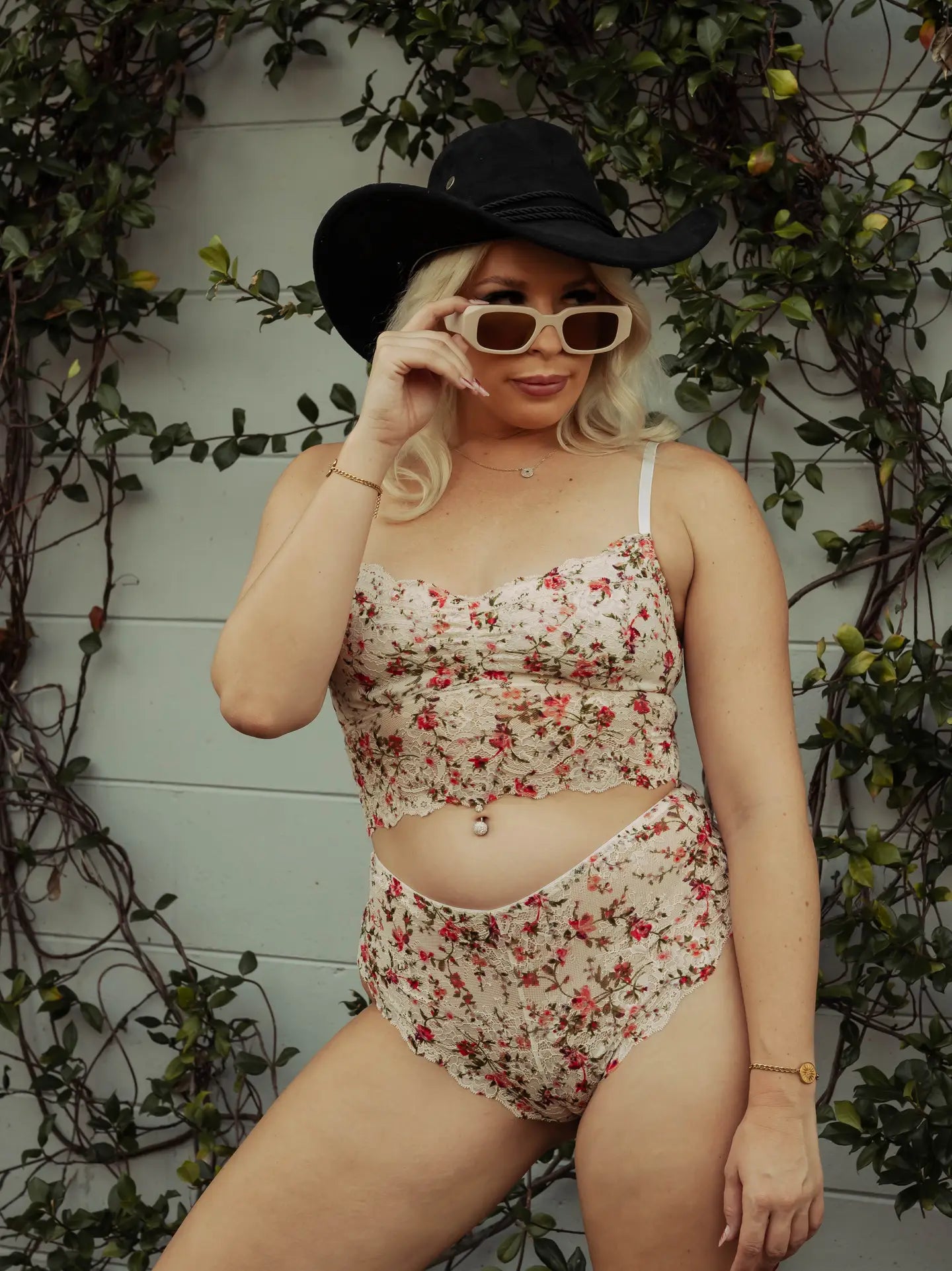 Woman wearing a floral bralette and underwear set with a cowboy hat and sunglasses, on a leafy background.