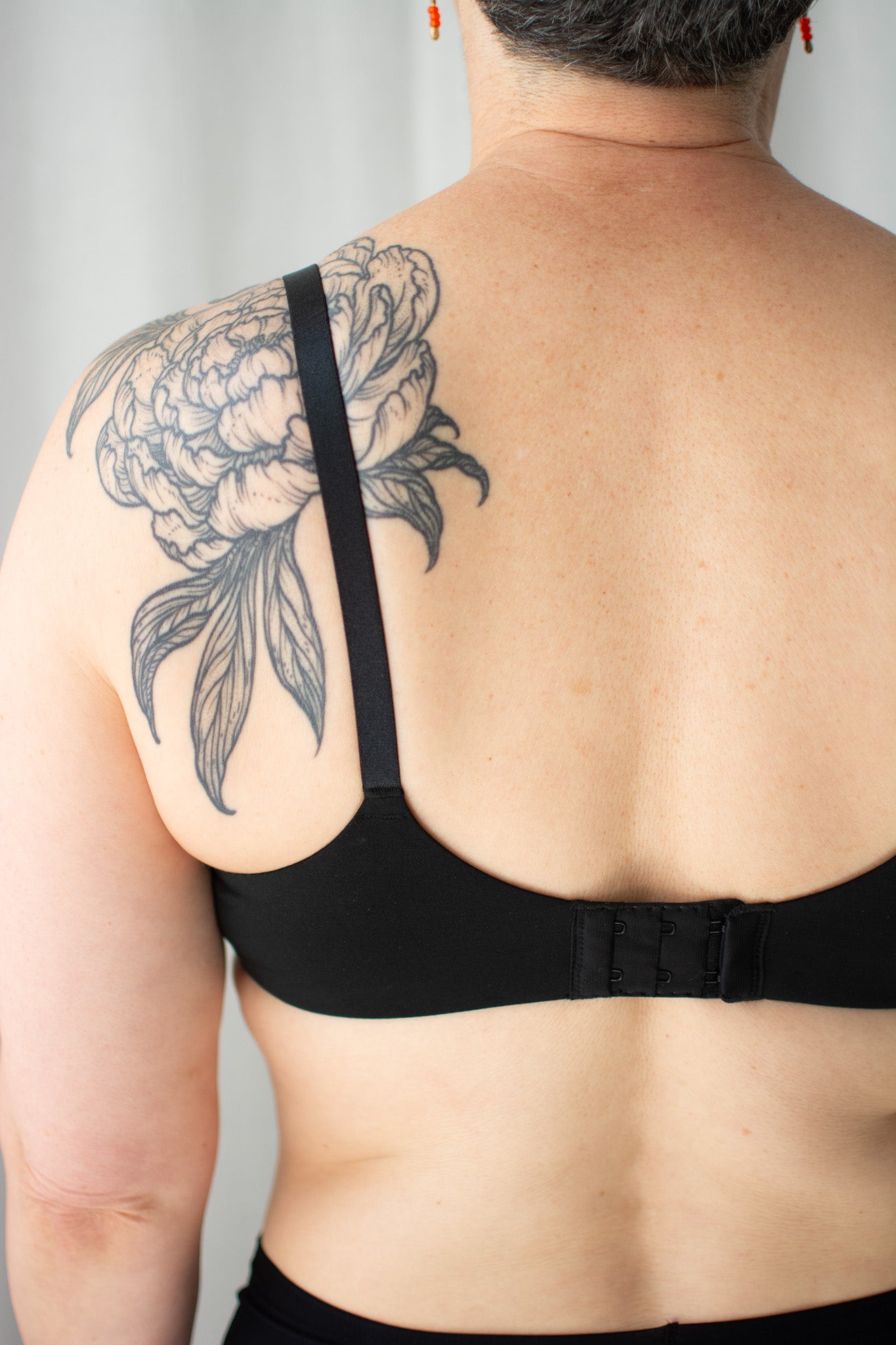 Person wearing a black bra with a floral tattoo on their shoulder against a plain background