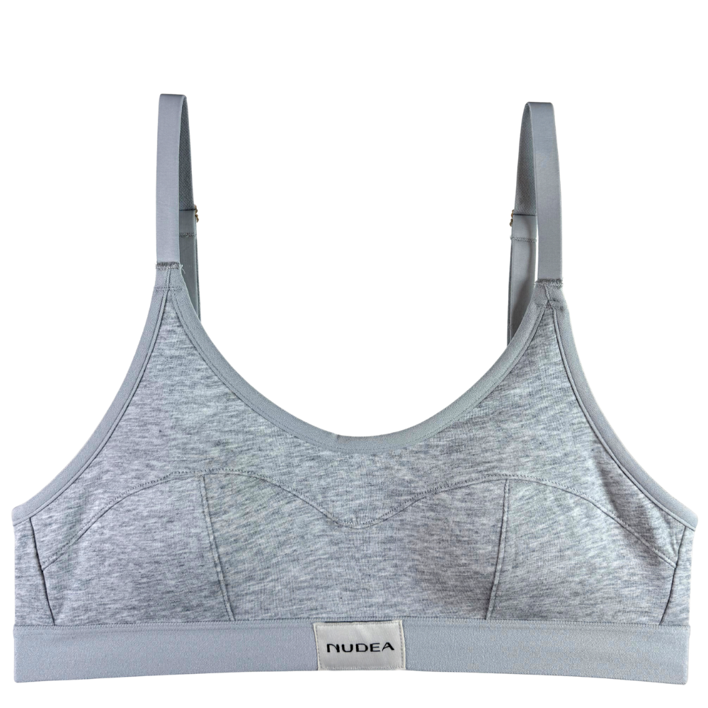 A light gray heathered sports bra with matching gray straps and a white brand label at the center bottom that reads NUDEA, shown against a plain white background.
