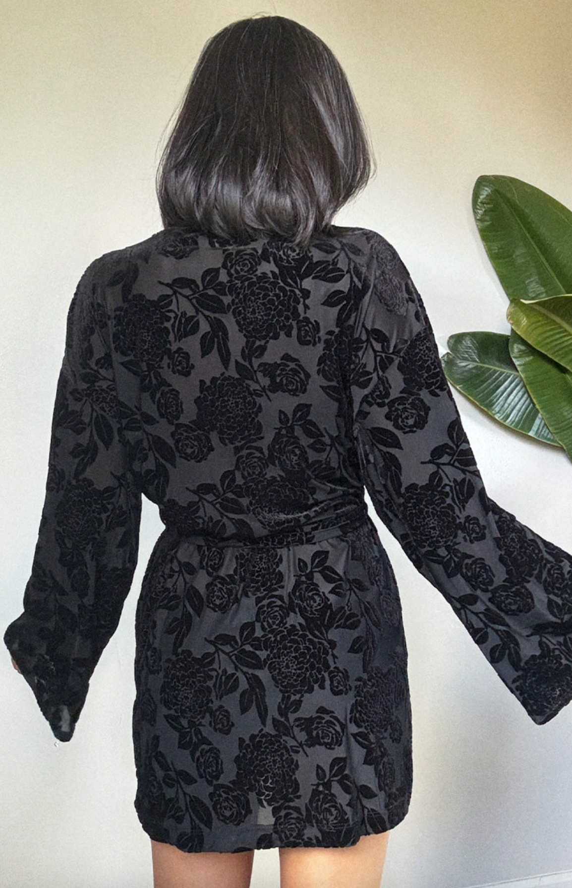 A woman viewed from behind wearing a black long-sleeve mini dress with a tonal floral jacquard pattern, standing against a light beige wall with a green plant leaf visible on the right.