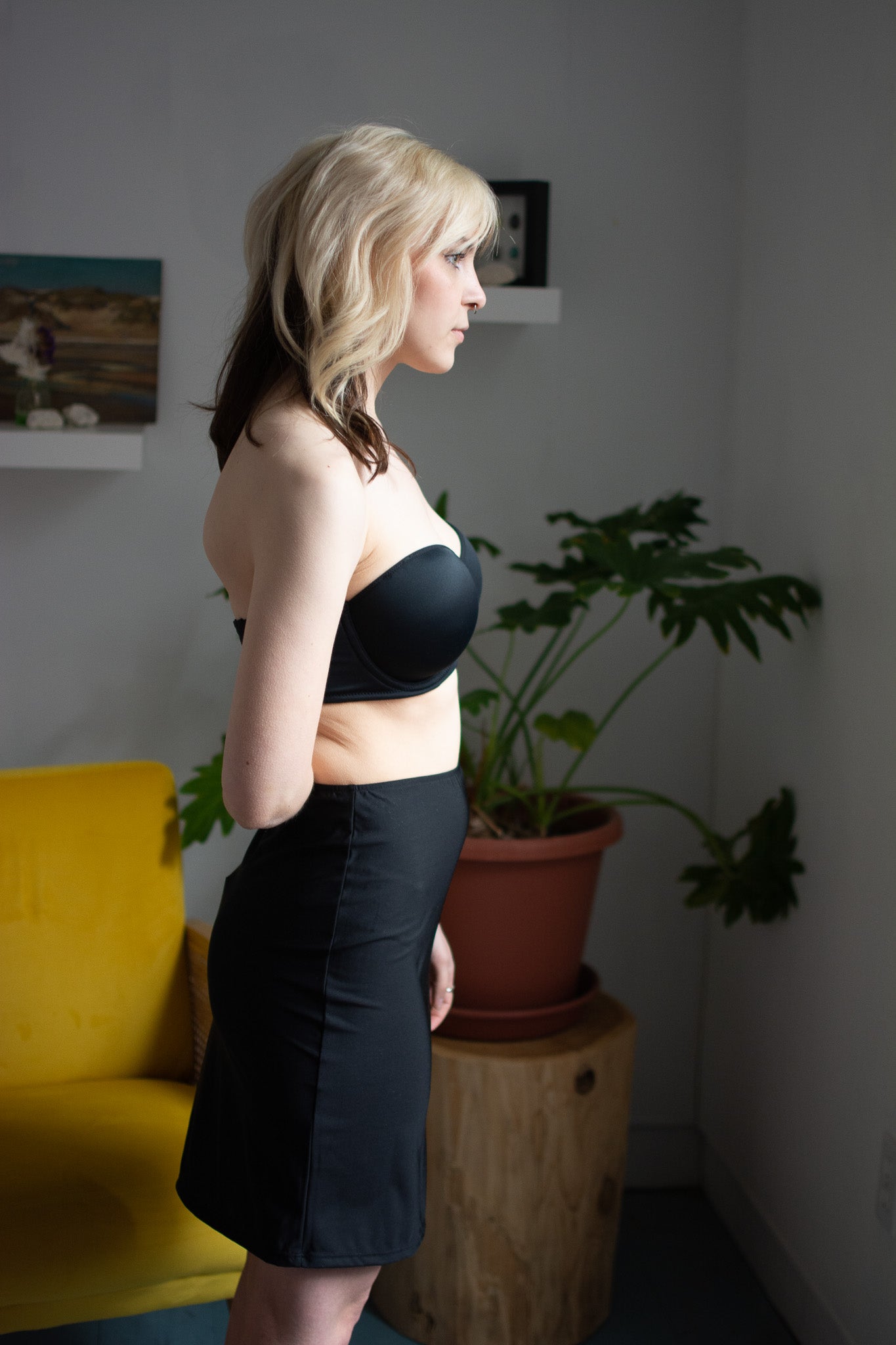 Woman in a black strapless top and black shorts standing in a room with a yellow chair and potted plant.