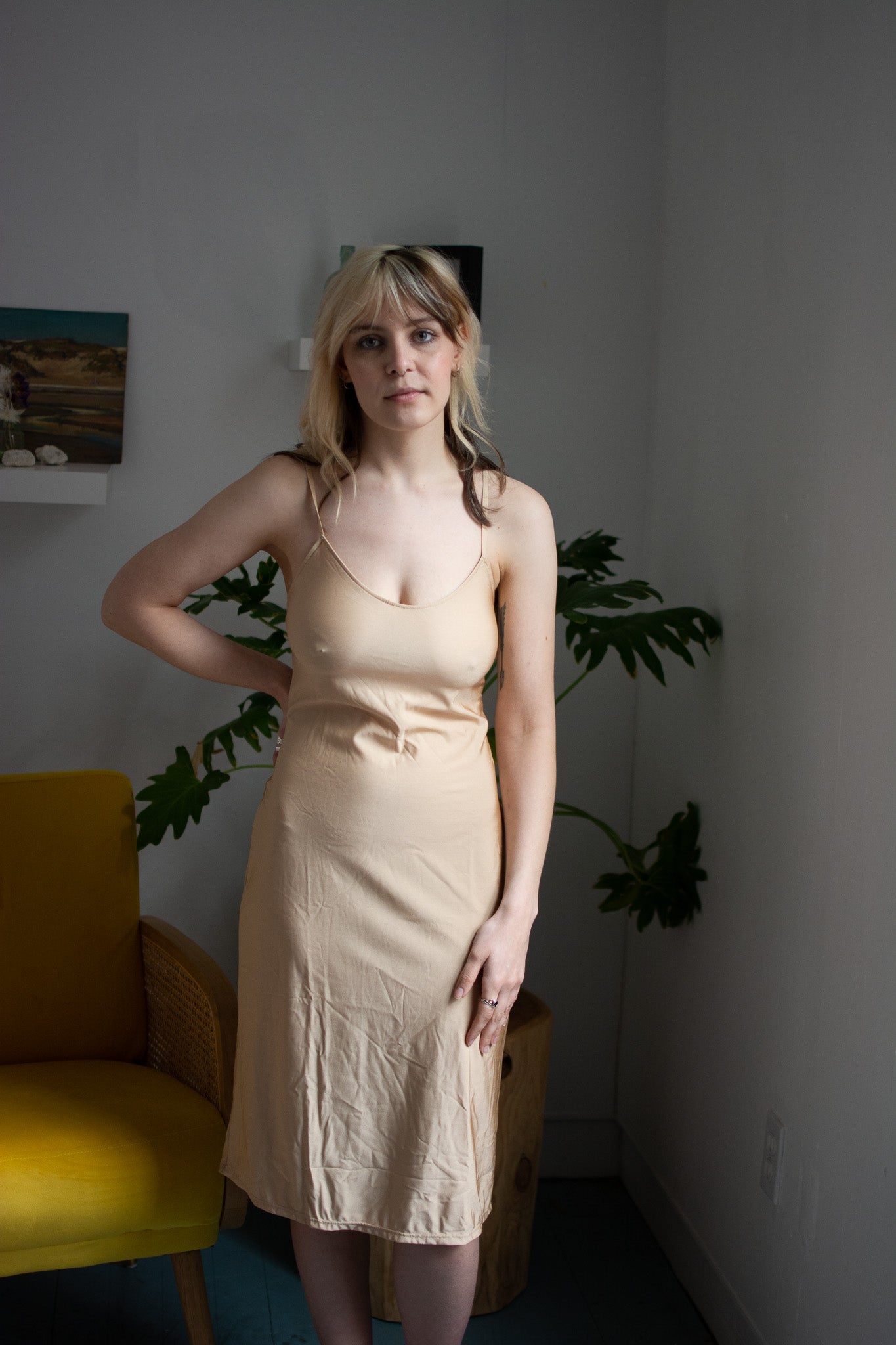 Woman wearing a beige dress standing in a room with a yellow chair and plants.
