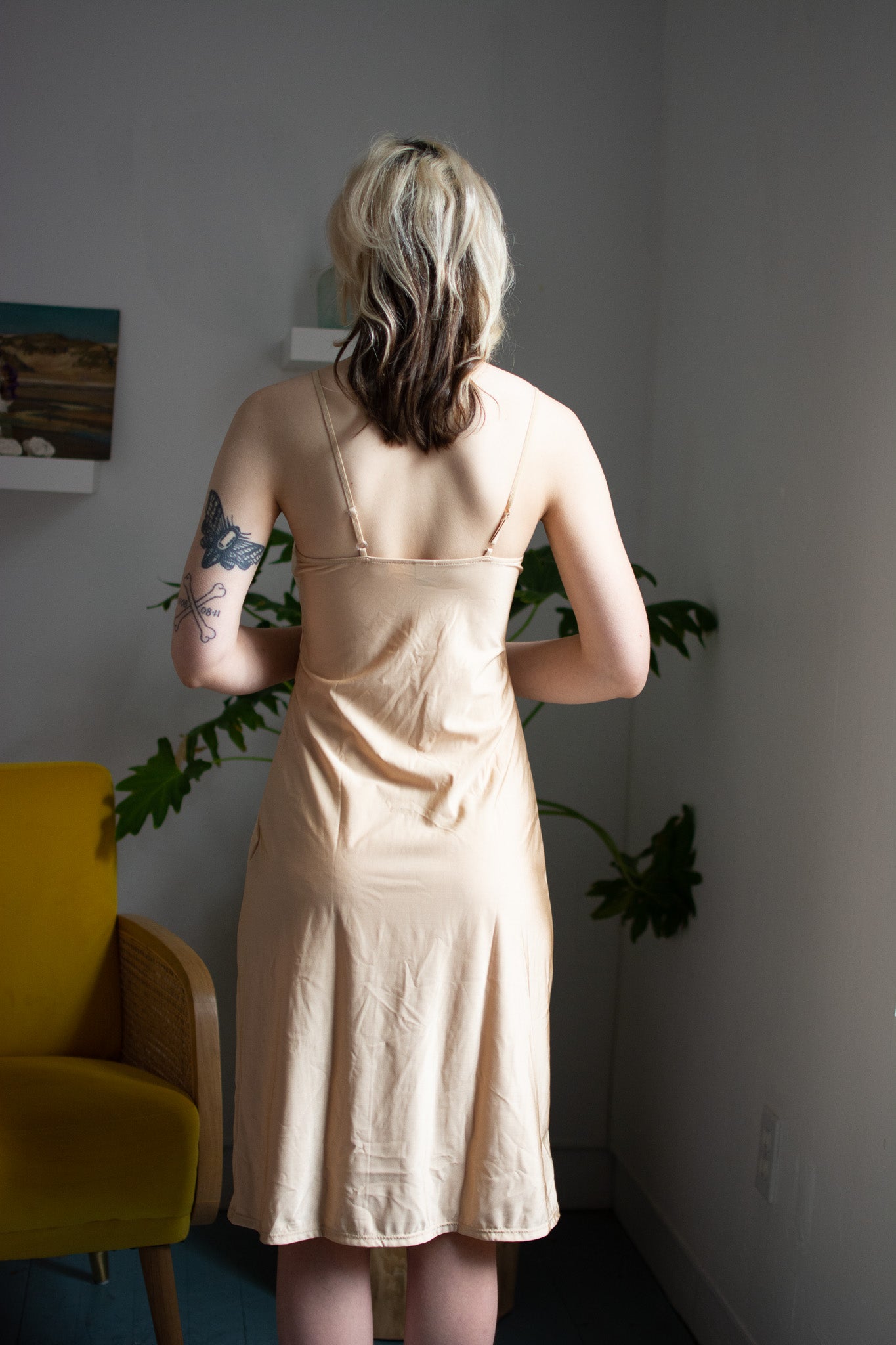 Person wearing a beige dress standing in a room with a yellow chair and plants.
