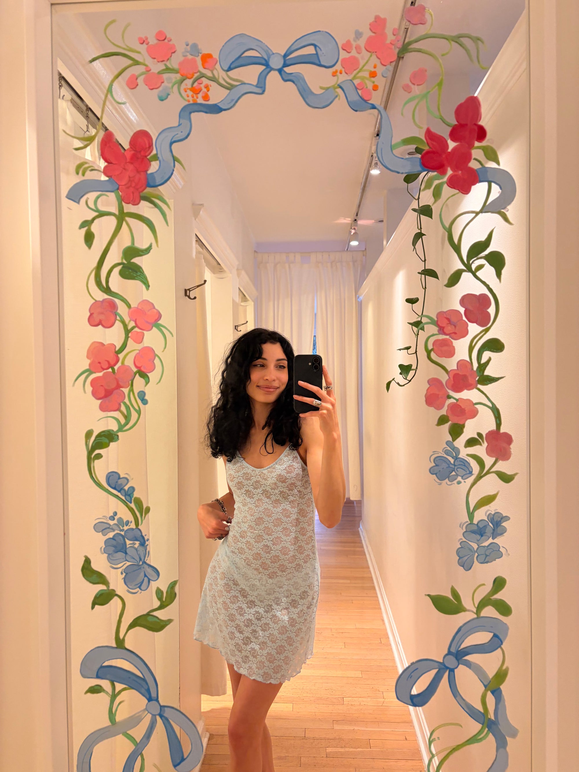 Woman taking a mirror selfie in a room with decorative floral and ribbon designs on the wall.