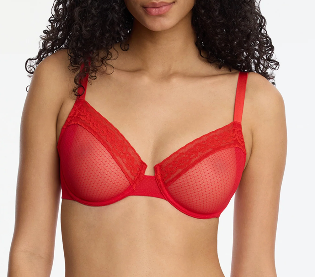 A close-up of a model wearing a bright red wireless bra with sheer mesh cups and lace trim, set against a plain light gray background.