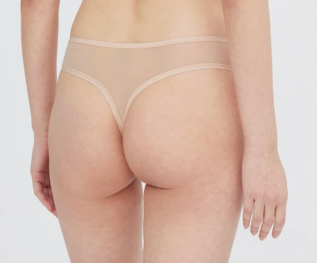 Rear view of a model wearing sheer nude beige mesh thong underwear with beige elastic bands, set against a plain light gray background.