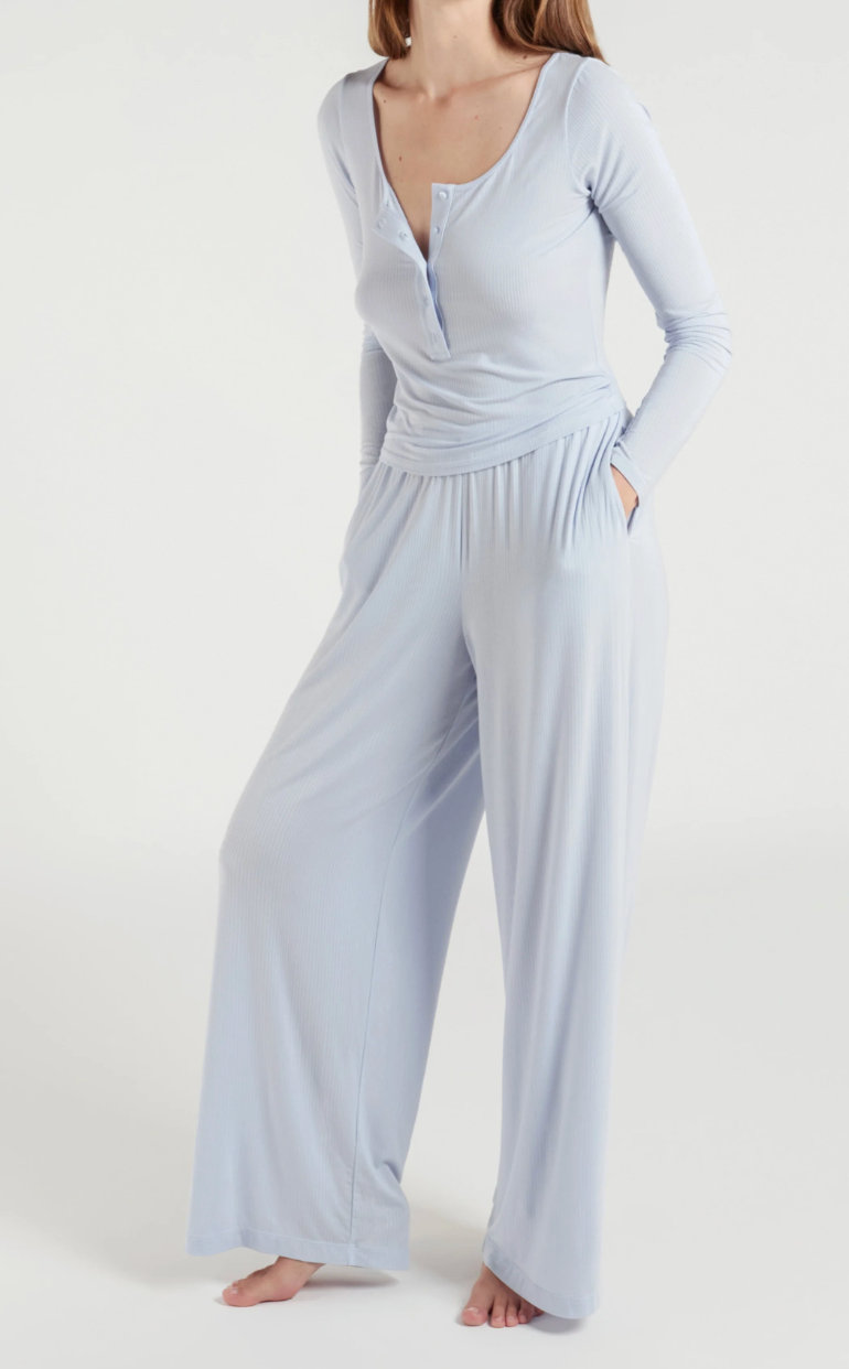 A woman modeling a light blue long-sleeve jumpsuit with a draped henley-style top and wide-leg pants, photographed against a plain light gray studio background.