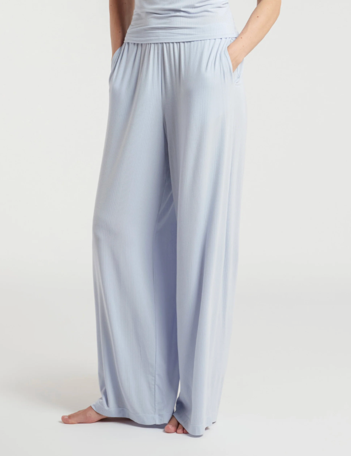 A person wearing light blue wide-leg lounge pants with a gathered elastic waistband and side pockets, shown from the torso down against a plain light gray studio background.
