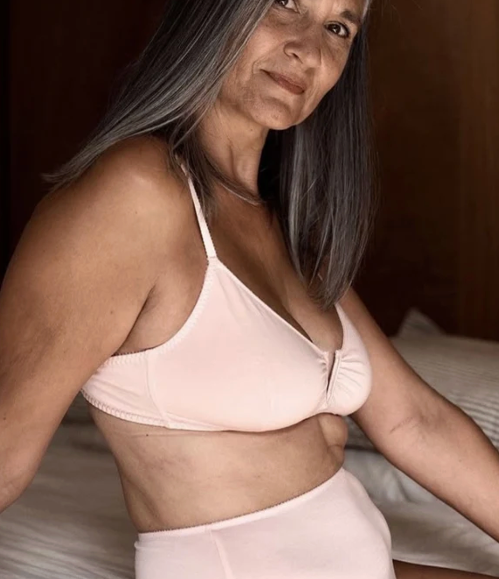 A woman with gray hair wearing a light pink two-piece lingerie set consisting of a bralette and matching high-waisted briefs, posed in a bedroom setting.