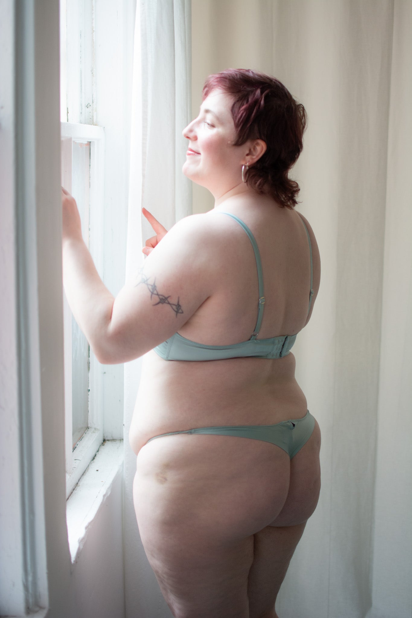 Woman in light blue lingerie standing by a window with white curtains.