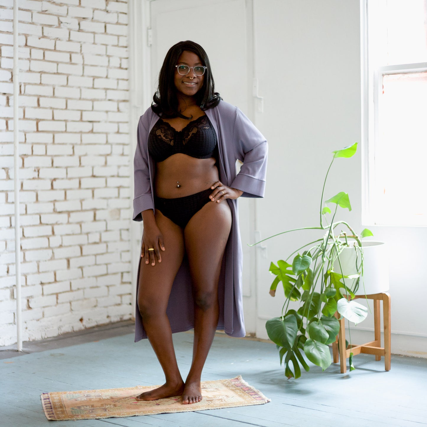 Model wearing black lace full cup bra, matching underwear, and a lilac robe in a sunlit room. 